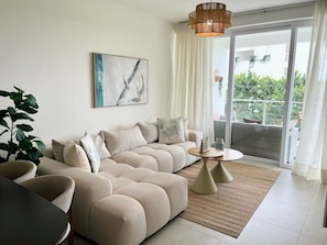 A modern living room with a sectional sofa, a small round table, and a large window with a view of greenery. - Stylish 2BR Condo with Pool, BBQ and Tropical Views in Punta Cana (Punta Cana)