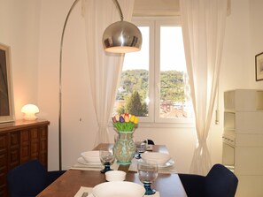 Ein Essbereich mit Tisch für zwei Personen, einer Blumenvase, einer Lampe und einem Fenster mit Blick auf eine Hügel.