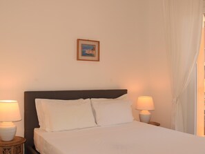Ein Bett mit weißer Bettwäsche, zwei Nachttischlampen, ein gerahmtes Bild an der Wand und ein Fenster mit Vorhängen.