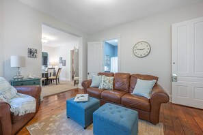 A living room with a brown leather sofa, two blue ottomans, a dining area with a table and chairs, and a clock on the wall.