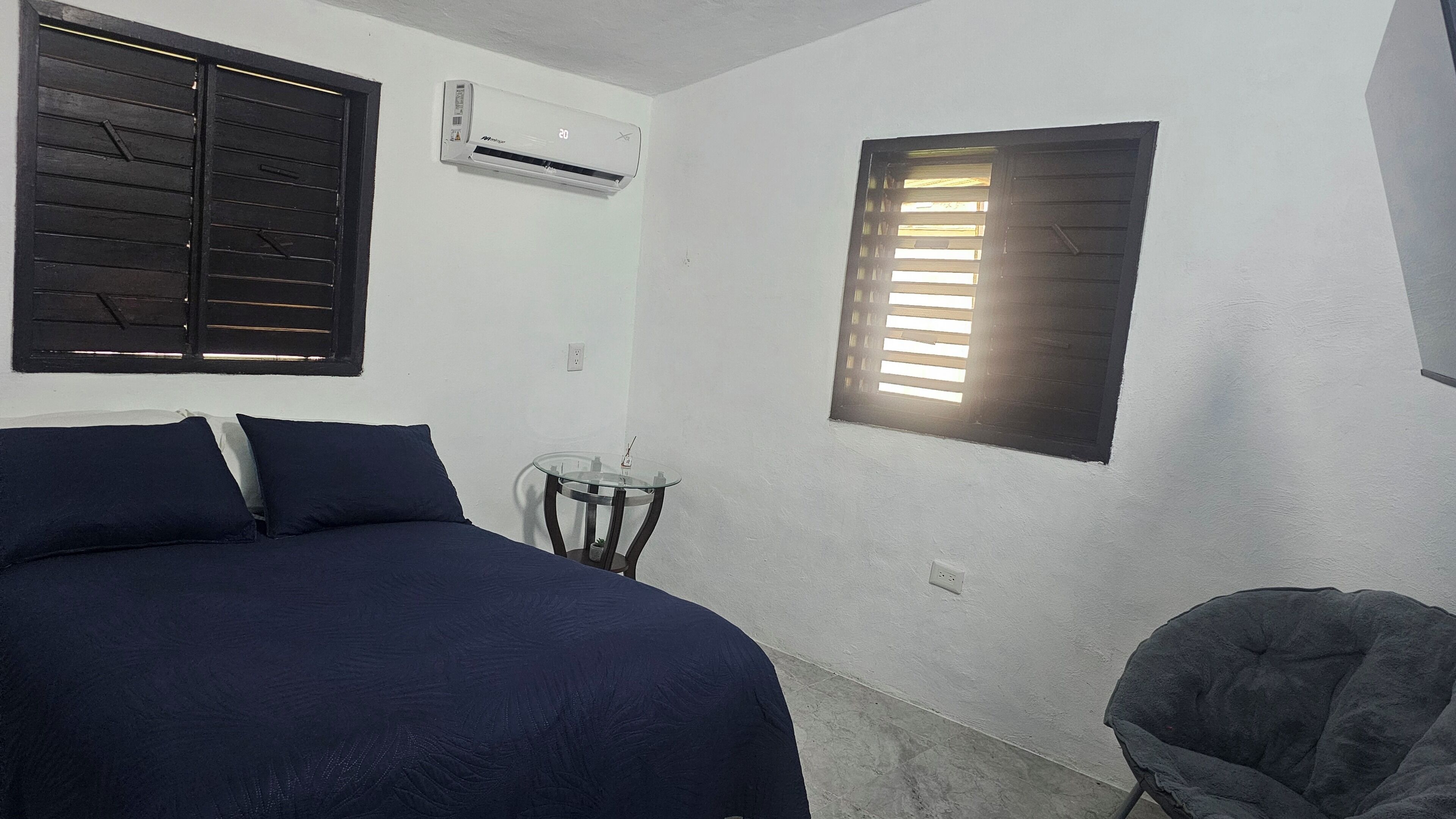 Quarto com cama, uma pequena mesa, cadeira, ar condicionado e janela com persianas.