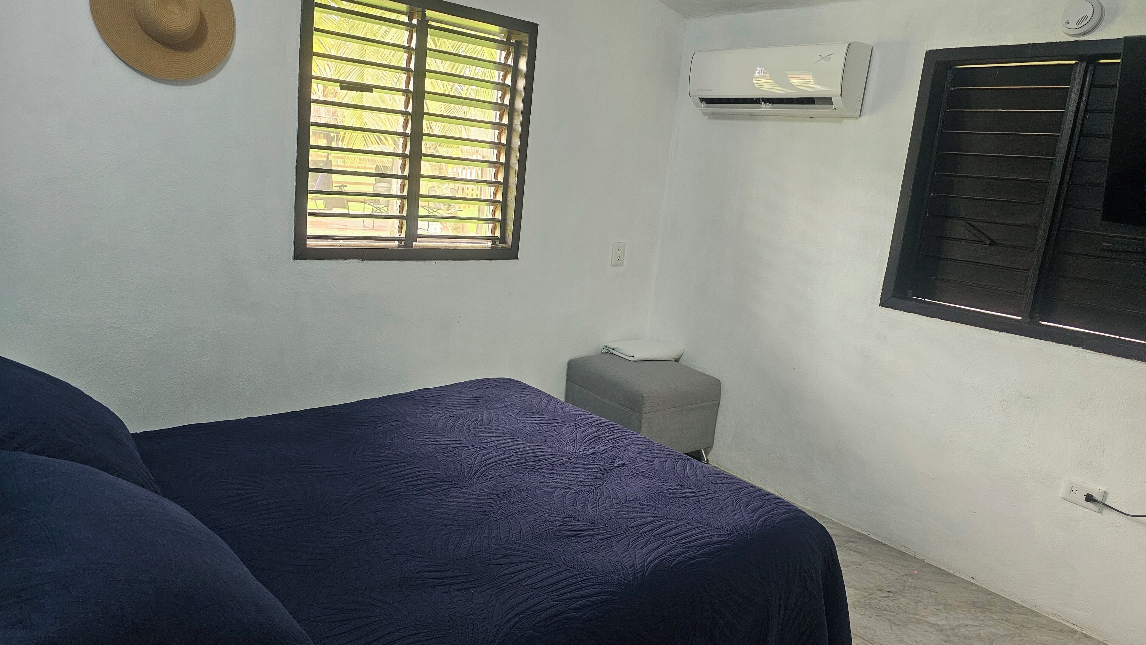 A room with a bed, a window with blinds, an air conditioning unit, and a small stool.