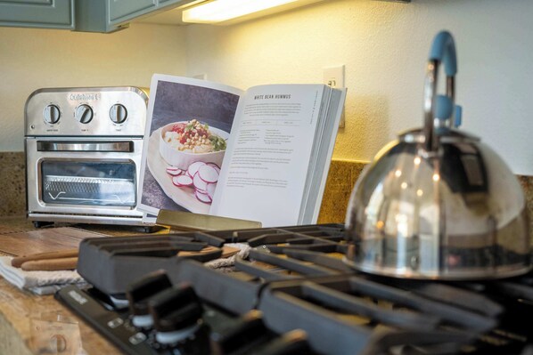 Una cocina con horno Cuisinart, un libro de cocina abierto en una página de receta y una estufa a gas con un hervidor.