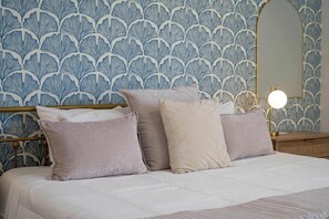 Un lit avec une couette blanche et deux oreillers, une table de chevet avec une lampe, et un mur orné d’un motif de feuilles.