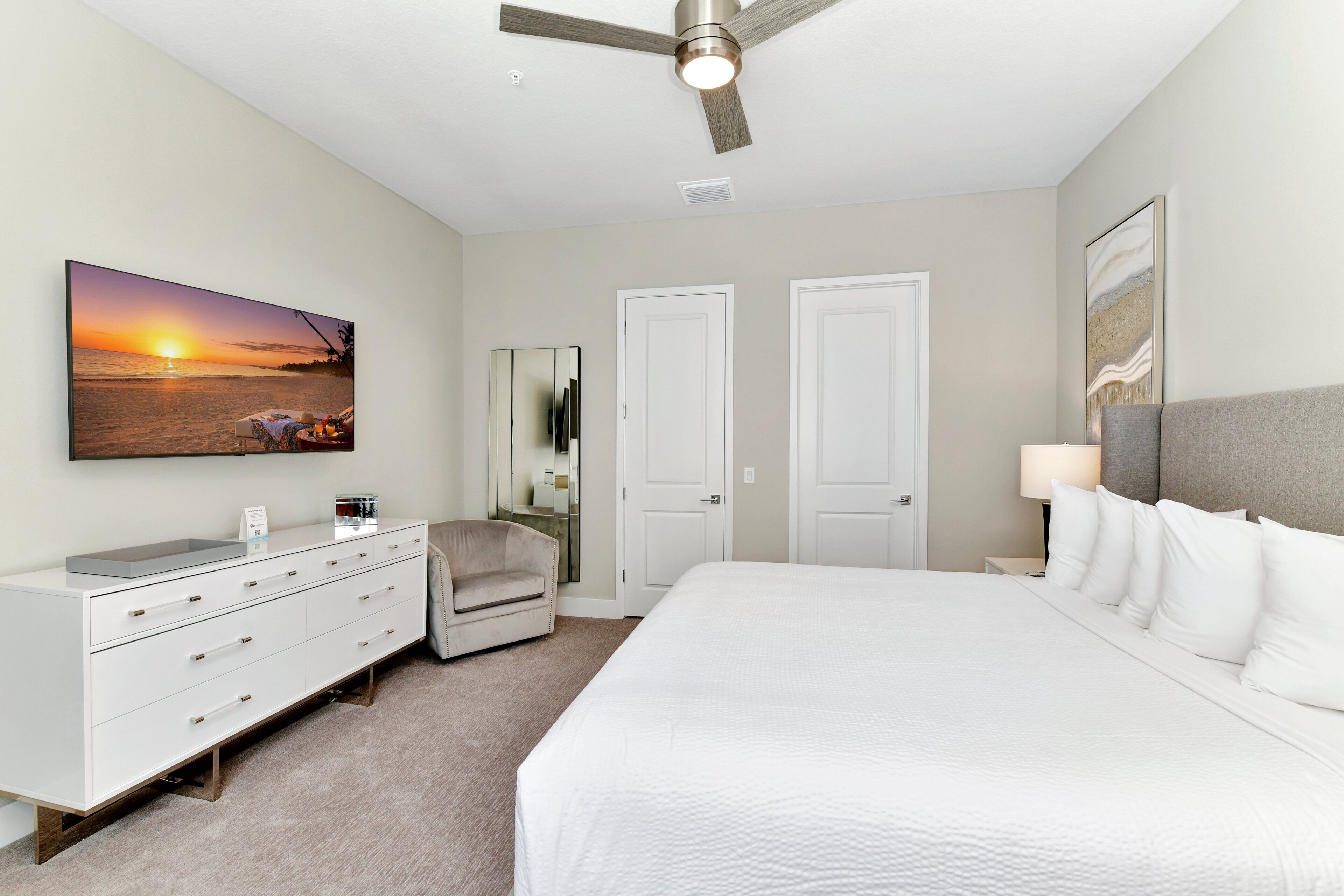 A bedroom with a bed, a dresser, a chair, a ceiling fan, and a painting of a sunset.