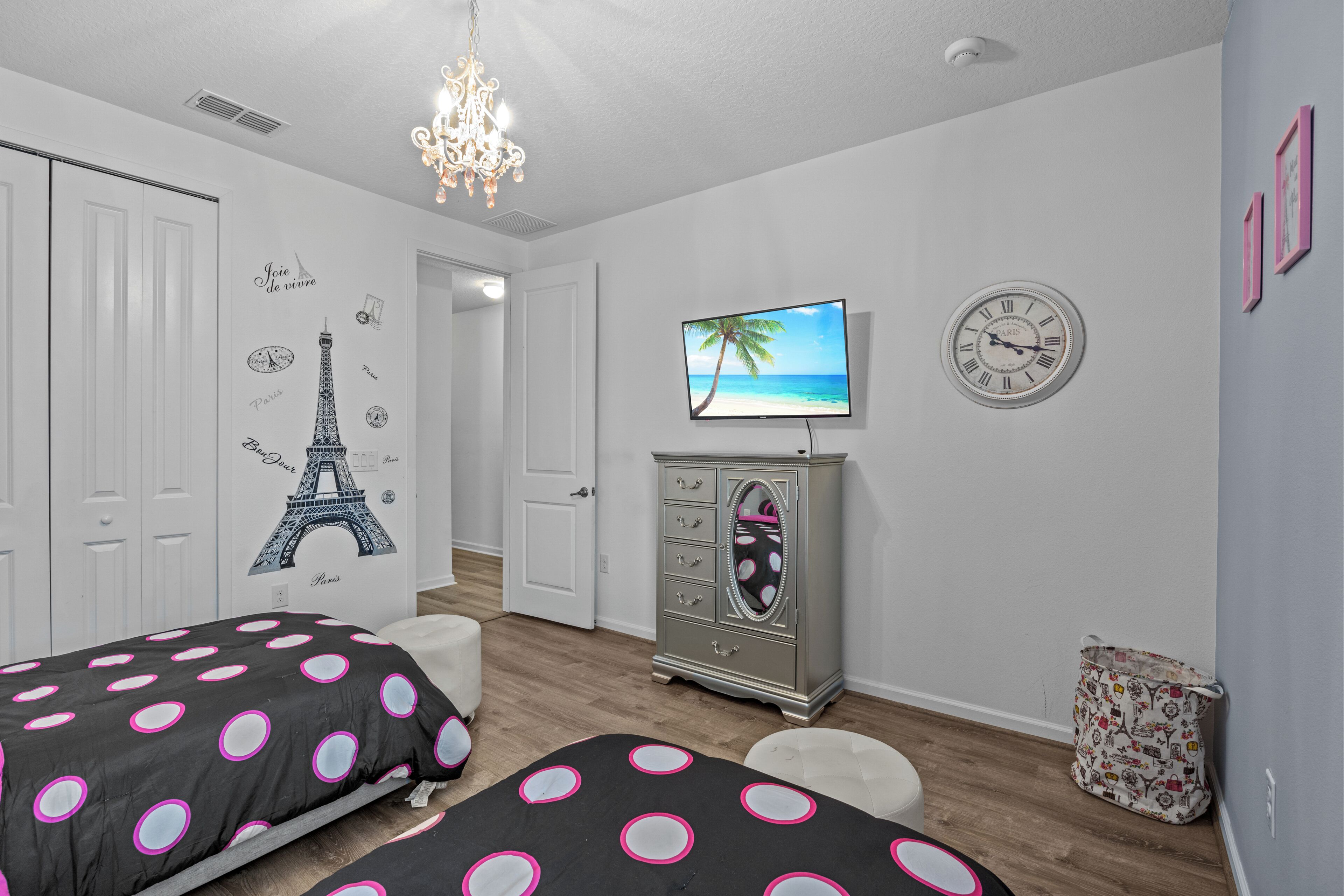 A bedroom with two beds, a dresser, a television, a clock, and a wall with Eiffel Tower decals.