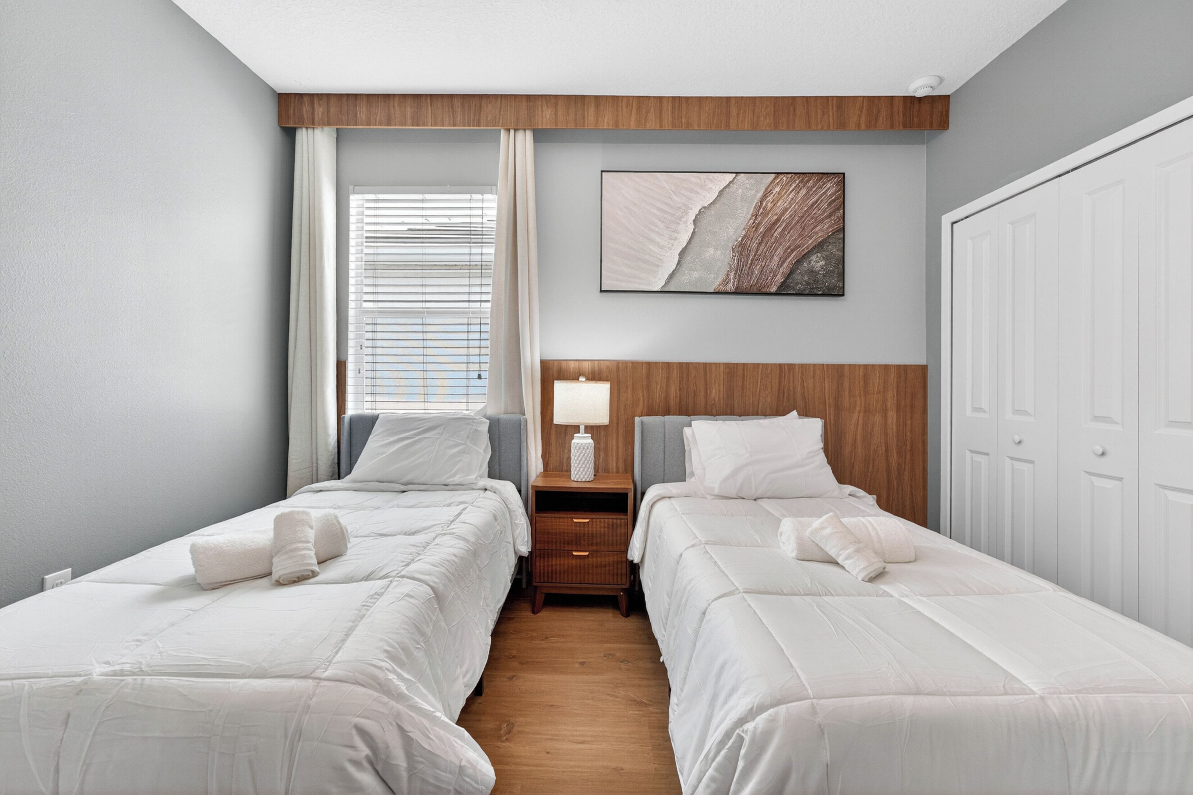 Une chambre d’hôtel avec deux lits, une tête de lit en bois, une table de chevet avec une lampe, un tableau au mur et une fenêtre avec des stores.