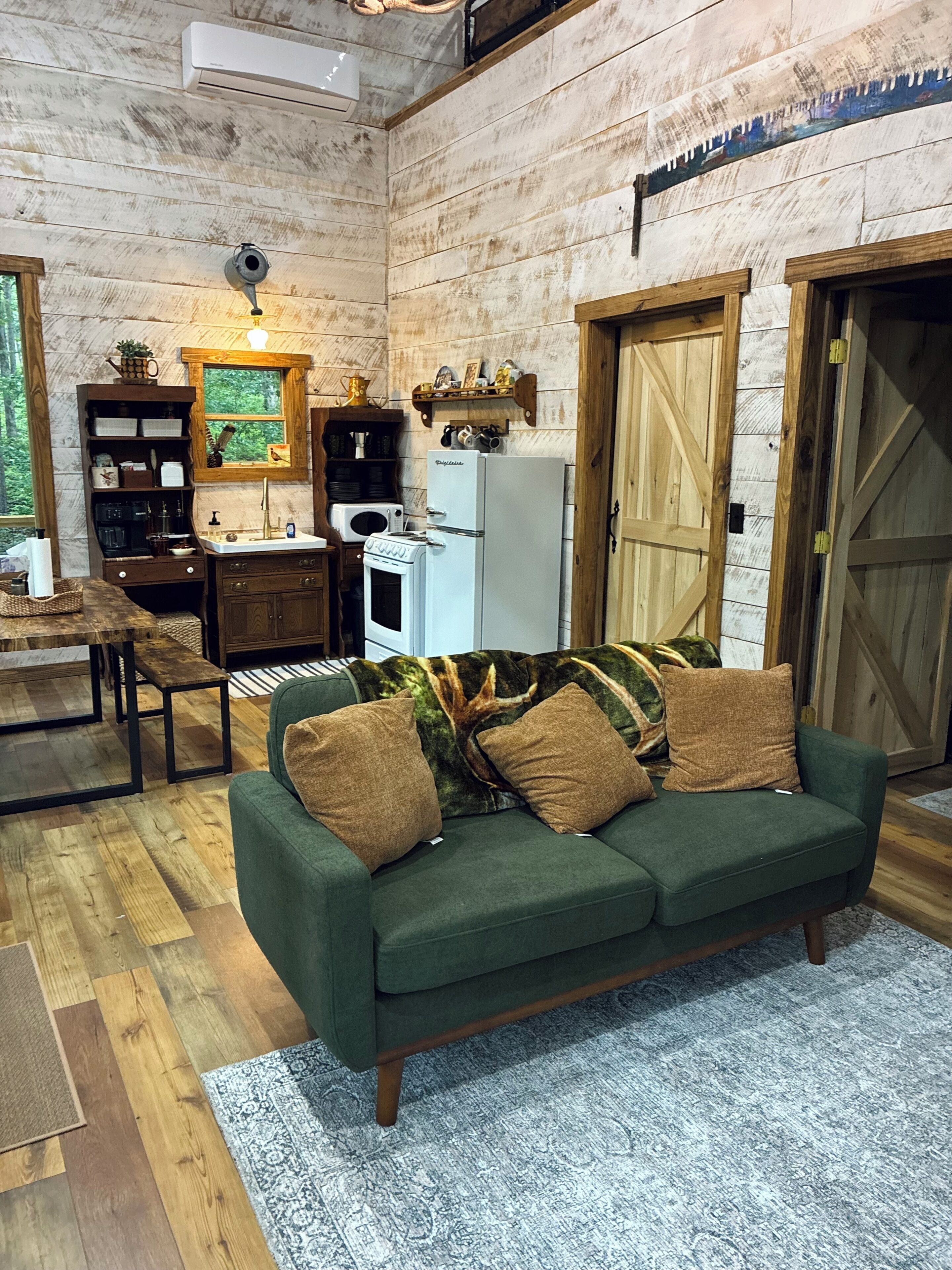 Een gezellig interieur van een chalet met een groene bank, houten vloeren, een keuken met een koelkast en een magnetron, en een grote houten deur.