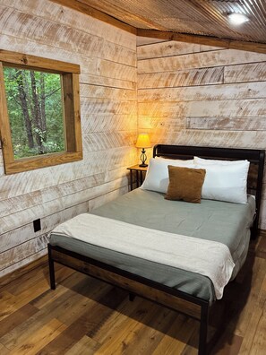 Ein Schlafzimmer mit Holzwänden, einem Bett, einem Nachttisch, einer Lampe und einem Fenster mit Blick auf einen Wald.