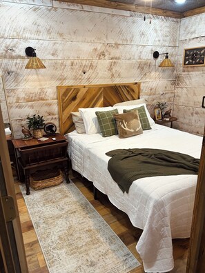 Une chambre à coucher avec une tête de lit en bois, un lit recouvert d’une literie blanche, un plaid vert, une table de chevet avec une lampe et une petite table ornée d’objets décoratifs.