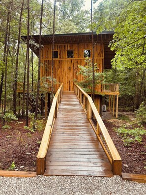 A wooden cabin with a wooden deck leading to a door, surrounded by a forest.