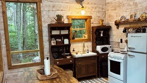 A rustic kitchen with wooden walls, a window with a forest view, a stove, a refrigerator, and a wooden table with benches. - Treehouse+Quiet Escape+National Forest+Firepit (Falkner)