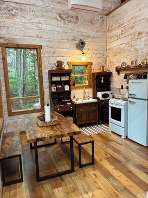 A rustic kitchen with wooden walls, a window with a forest view, a stove, a refrigerator, and a wooden table with benches.