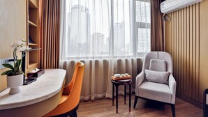 A hotel room with a curved marble desk, a chair, a small table with a tray, a potted plant, a window with curtains, and an air conditioning unit.