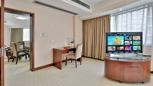 A hotel room with a desk, chair, TV, and a large window with curtains.