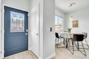 Una habitación con puerta azul, pared blanca, mesa de comedor con sillas, lámpara colgante y un cuadro enmarcado.