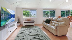 A living room with a flat-screen TV, a beige sofa, a white entertainment unit, and a large window with a view of greenery. - Ocean Soul Unit 1 (Burnett Heads)