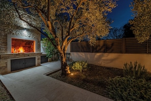 A stone fireplace with a fire, surrounded by a garden with plants and a tree. - Olive House Pool & Garden, Zemunik Donji, Croatia (Zemunik Donji)