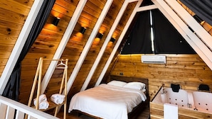 A triangular attic bedroom with wooden beams, a bed, a hanging chair, and a small window.