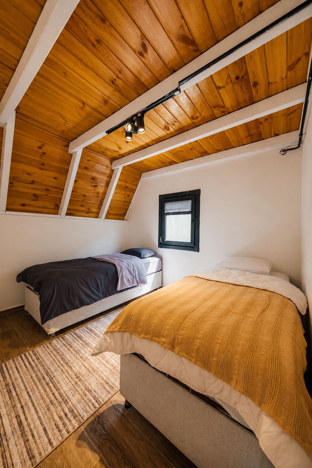 A bedroom with a sloped ceiling, two beds, a window, and a wooden floor.