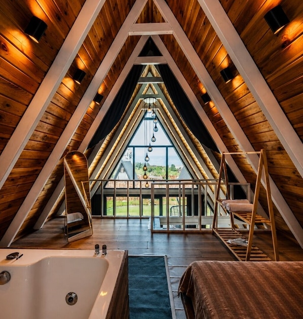 A triangular attic bedroom with a large window, a bathtub, and a bed.