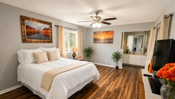 A bedroom with a bed, a ceiling fan, a window with curtains, a painting, a mirror, and a TV.