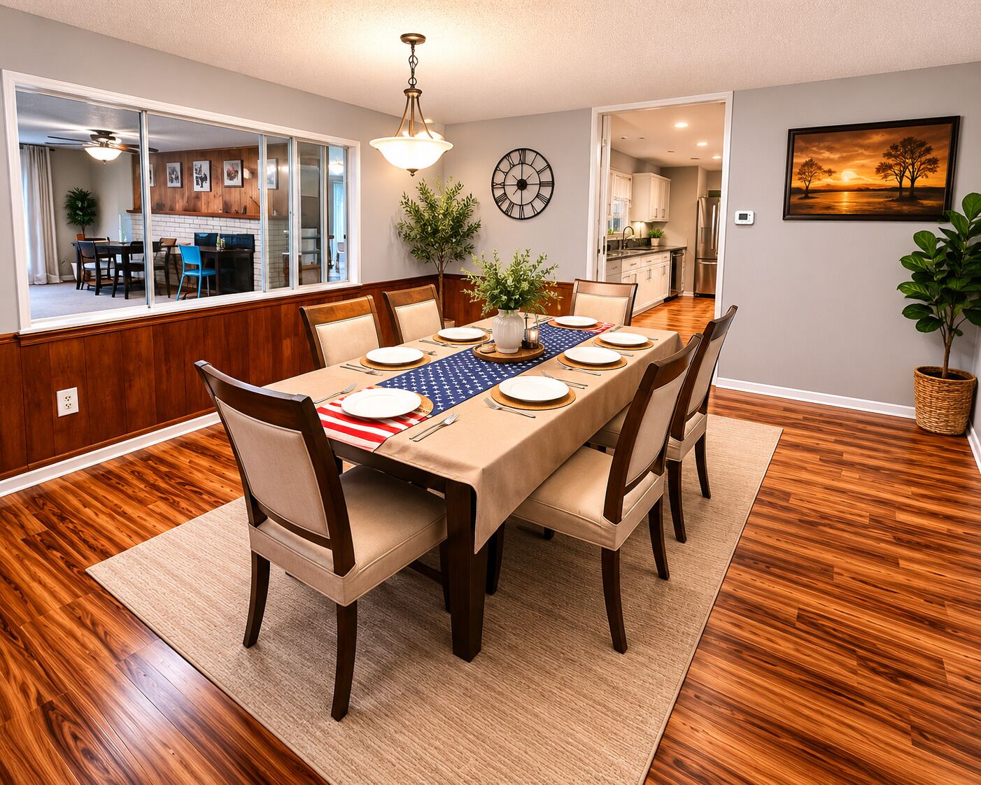 Une salle à manger avec une table en bois dressée pour six personnes, une horloge au mur et une cuisine en arrière-plan.