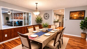 A dining room with a wooden table set for six, a clock on the wall, and a kitchen in the background.
