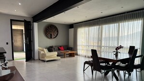 En moderne stue med sofa, sofabord, spisebord og stole. Der er et stort vindue med lette gardiner, en dør til udendørs og et billedværk hængt på væggen.