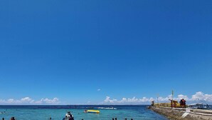 A beach with clear blue water, people swimming, and a lifeguard on duty. - Huge Lux. 2 BR Condo (100 SQM)  2 pools - Beach, Sea-views, Gym, Tours available (Lapu-Lapu City)