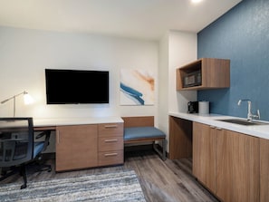 Ein kleines Zimmer mit Schreibtisch, Stuhl, Fernseher, Mikrowelle und Spüle.