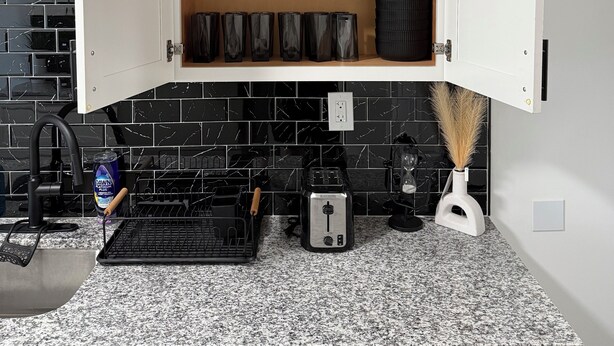 Una cocina con un armario blanco, baldosas negras, encimera de granito, un tostador y un lavabo.