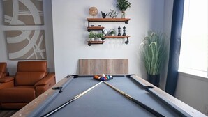 Una mesa de billar con un cabecero de madera, un sillón de cuero marrón y una estantería con plantas y objetos decorativos.