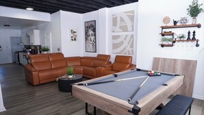 Una sala de estar moderna con mesa de billar, sofá de cuero marrón y cocina al fondo.