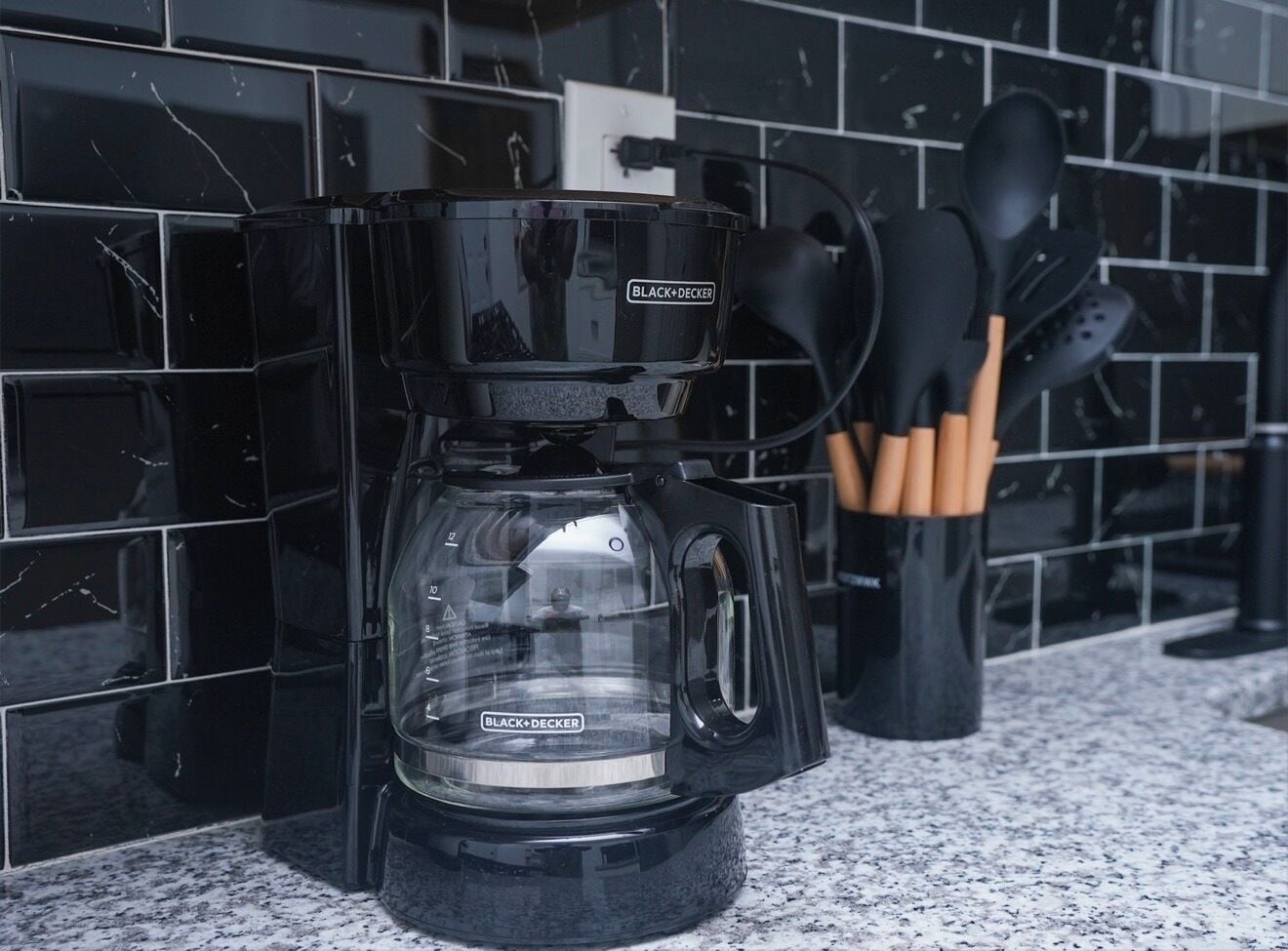 Una cafetera BLACK+DECKER sobre una encimera de granito con un salpicadero de azulejos negros.