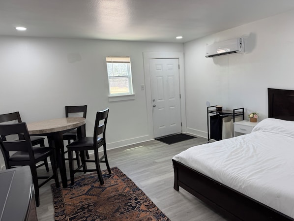 A bedroom with a bed, a desk with a chair, a small table with chairs, a window, and an air conditioning unit.