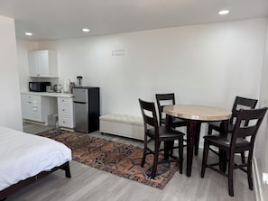 A compact room with a bed, a small kitchenette, a round dining table with chairs, and a patterned rug.