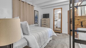A bedroom with a bed, a dresser, a television, and a bathroom visible through an open door. - Guesthouse in Mendenhall Valley (Juneau)