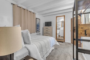 A bedroom with a bed, a dresser, a television, and a bathroom visible through an open door. - Guesthouse in Mendenhall Valley (Juneau)