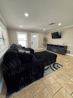 A living room with a sectional sofa, a flat-screen TV, and a large window with blinds.