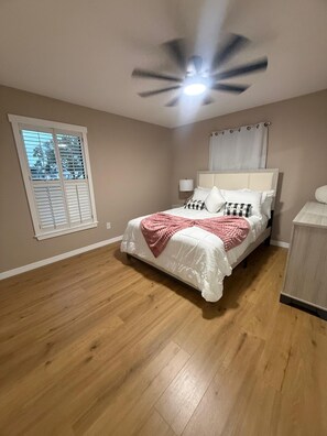 Une chambre à coucher avec un lit, un ventilateur de plafond, une fenêtre avec des stores et une table de chevet.