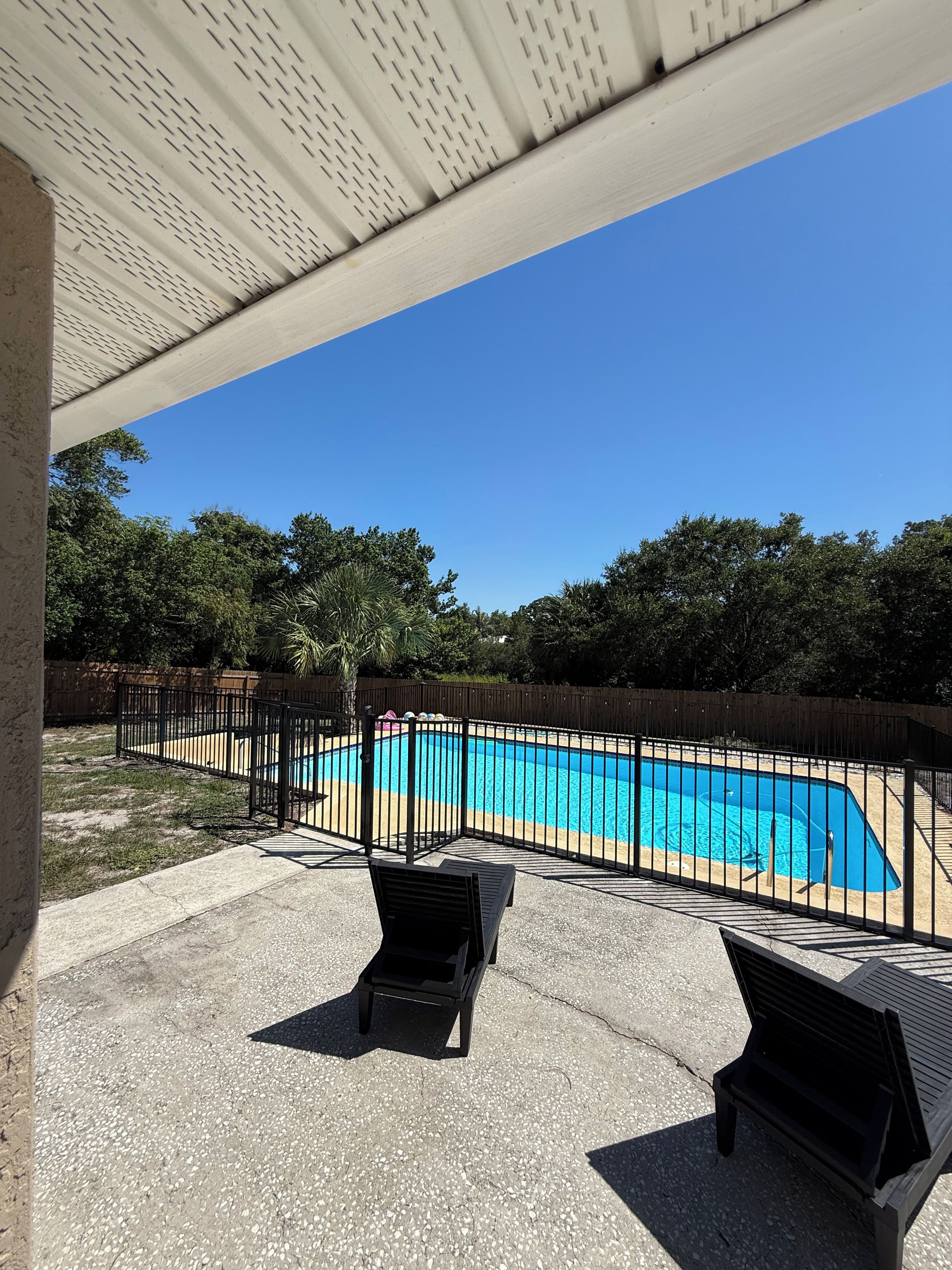 A swimming pool with a black metal fence, surrounded by trees and a wooden fence. There are two lounge chairs in the foreground.