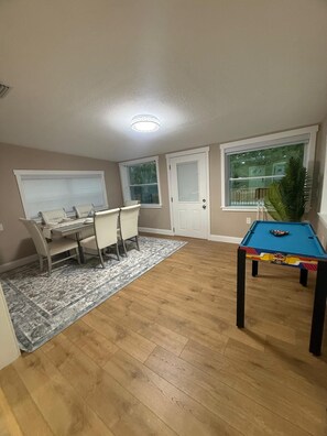 Una habitación con mesa de billar, una mesa de comedor con sillas, una ventana con persianas y una puerta.