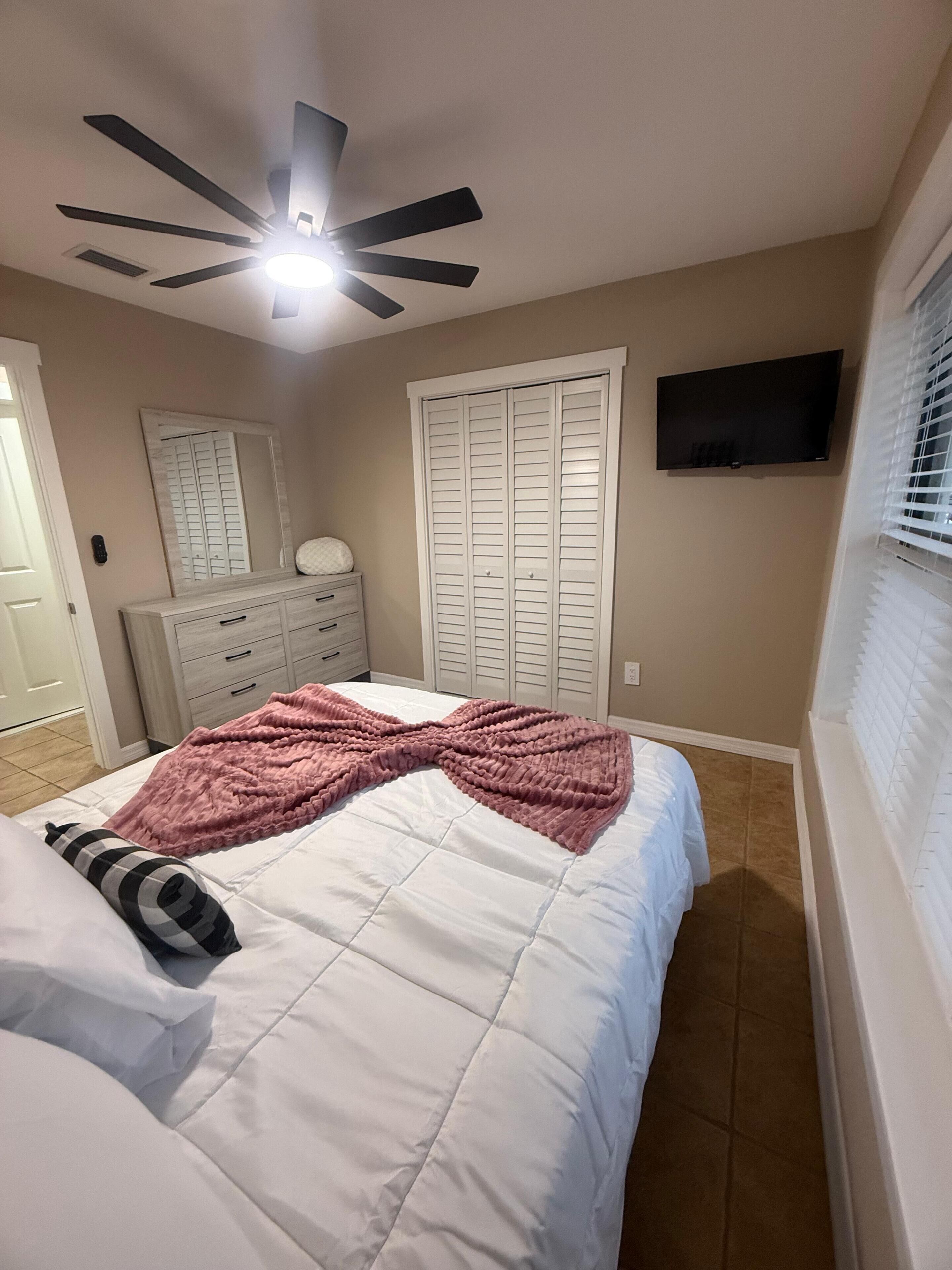 Un dormitorio con cama, ventilador de techo, televisión, armario y ventana con persianas.