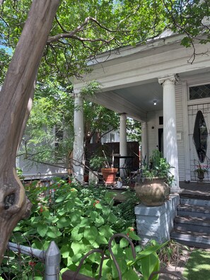 A covered porch with columns, potted plants, and a garden area with greenery and a tree. - The Artist's Room in an Antique Victorian House (Dallas)