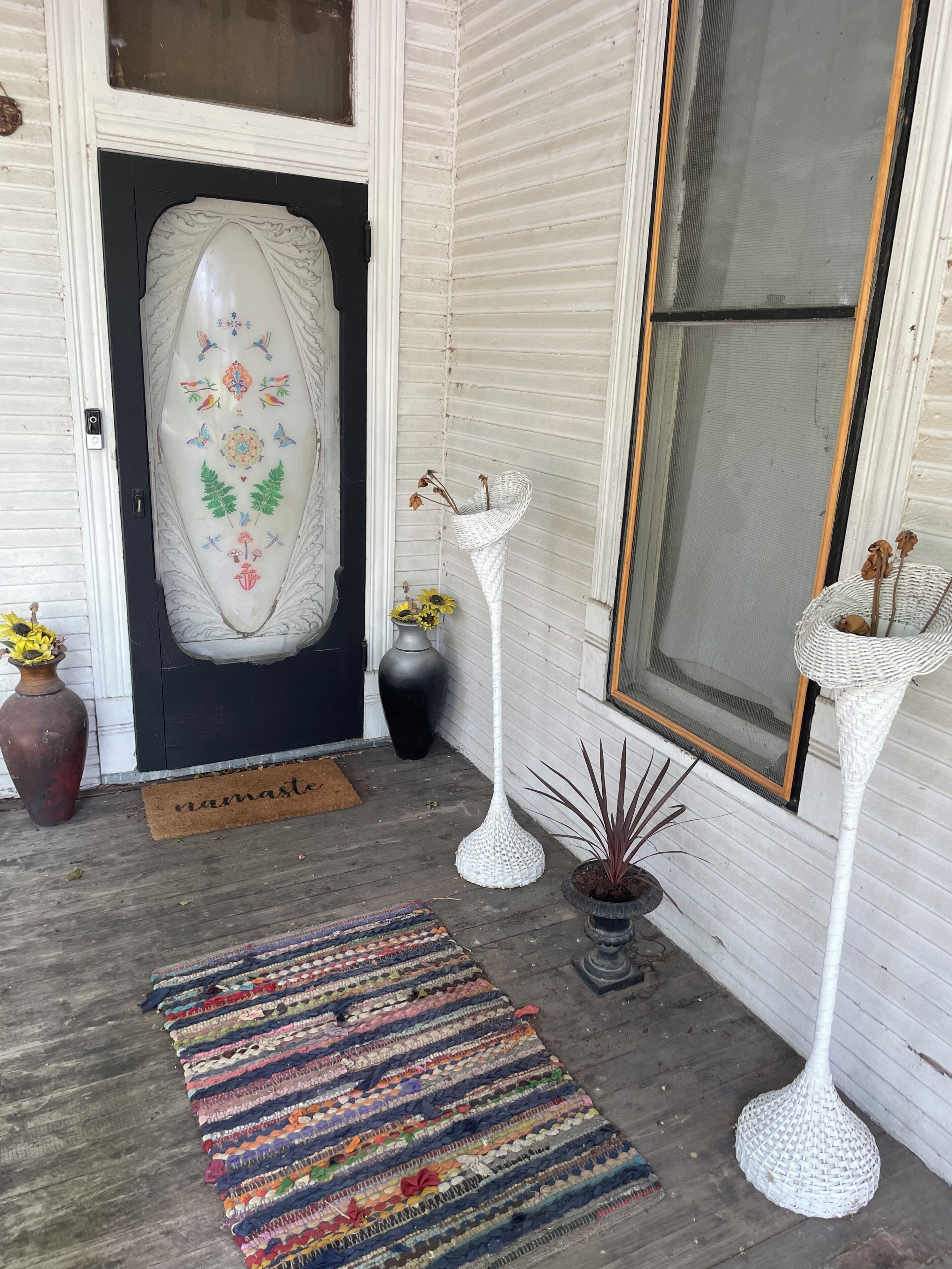 Un portico con una porta decorativa, una lampada da terra in vimini, un tappetino d'ingresso e un tappeto colorato.