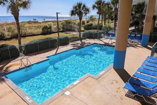 A swimming pool with clear blue water, surrounded by a fenced area with palm trees and a view of the ocean.