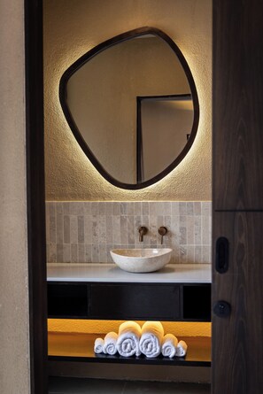 A bathroom with a round mirror, a white sink, and neatly rolled towels on a shelf. - SISI Wellness Villas (Pecatu)