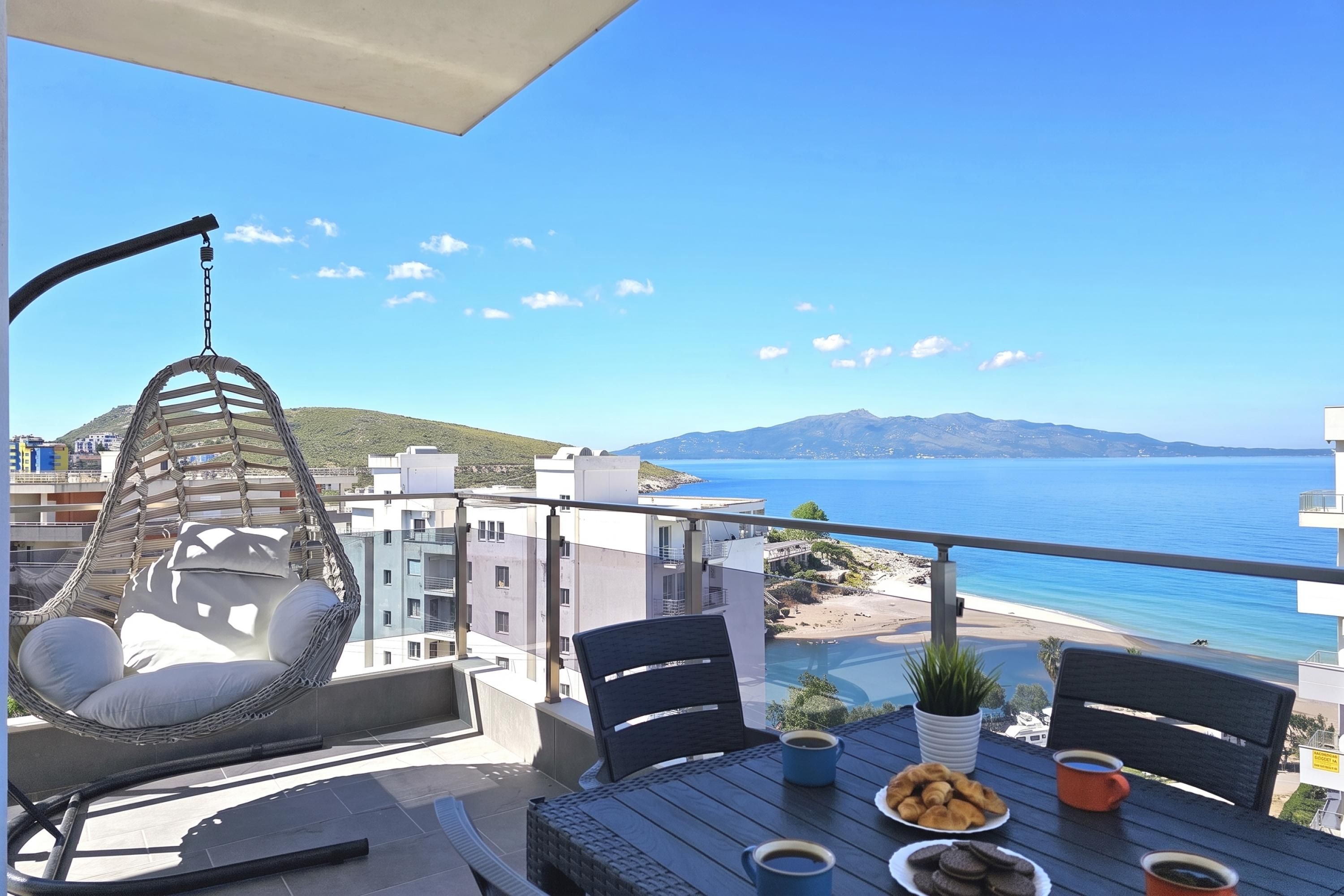 A balcony with a hanging chair, a table with coffee cups and pastries, and a view of the sea and distant mountains.