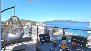 Ein Balkon mit einer Hängematte, einem Tisch mit Speisen und Getränken und Blick auf das Meer und Gebäude.